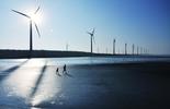 Turbinas eólicas em uma praia, com duas pessoas caminhando 