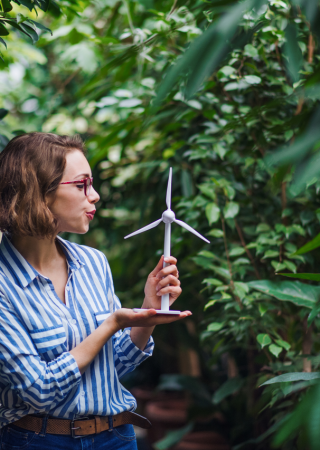 Imagem de uma mulher rodeada por folhas e segurando a miniatura de uma turbina de energia eólica